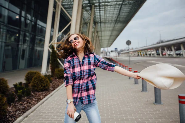 Молодая студентка-путешественница в клетчатой рубашке держит одноразовую чашку, прыгает и машет шляпой. Свобода путешествий и отпусков . — стоковое фото