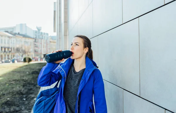 Joven corredora con bolsa de deporte bebiendo agua durante la carrera en la ciudad cerca de la pared . — Foto de Stock