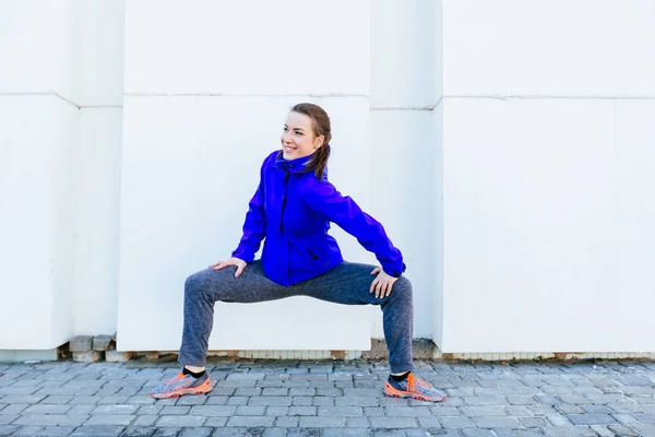 Joven atractiva mujer fitness en posición plie sobre fondo blanco de la pared en la calle . — Foto de Stock