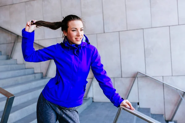 Mujer fitness en ropa deportiva azul corrigiendo el cabello mientras toma un descanso de correr relajante en los pasos . — Foto de Stock
