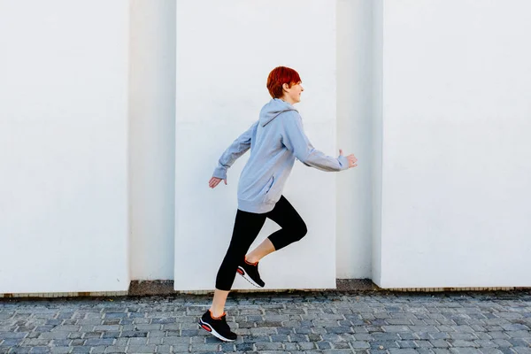 Νέοι κόκκινο κεφάλι σπορ γυναίκα που τρέχει κατά μήκος λευκό τοίχο. — Φωτογραφία Αρχείου