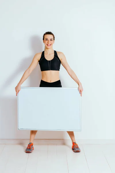Hermosa joven fitness mujer en chándal sosteniendo tablero en blanco aislado sobre fondo blanco — Foto de Stock