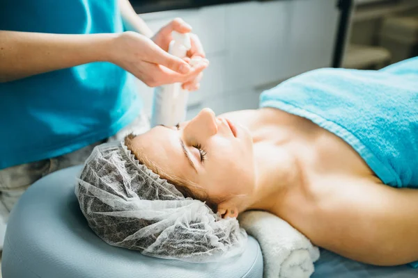 Hände der Kosmetikerin drücken die Körpercreme, die neben einer jungen Frau steht, die auf einem Massagetisch im Inneren eines gesunden Beauty-Spa-Salons liegt. — Stockfoto