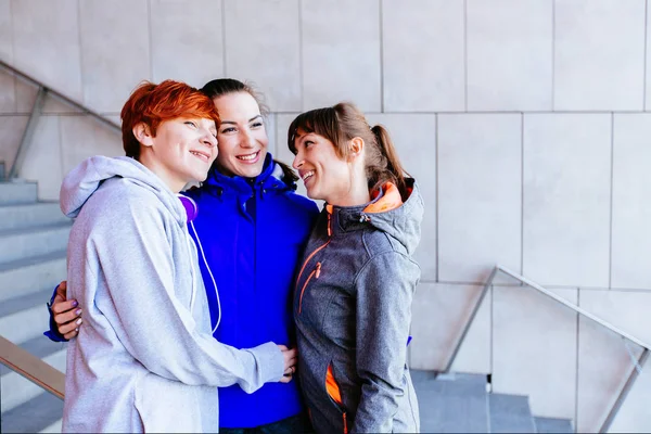 Un grupo de amigos, tres mujeres deportistas, se reúnen y se divierten en las escaleras de un edificio moderno. Vida sana, amistad y conceptos deportivos — Foto de Stock