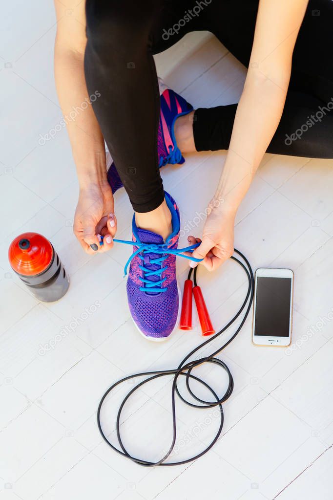 Top view of woman sitting on white wooden floor and tying her sport shoes. Skipping rope, bottle of water and mobile phone on the white wooden background. Sport, diet and healthy life concept.