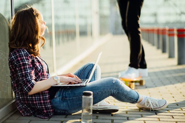 Счастливая молодая хипстерша сидит на полу и пользуется ноутбуком на сером городском фоне. Спорт, приключения и отдых . — стоковое фото