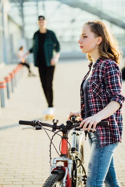 Молодая хипстерша в клетчатой рубашке, стоящая с велосипедом, ожидая своего парня на скейтборде. Концепция активного образа жизни — стоковое фото