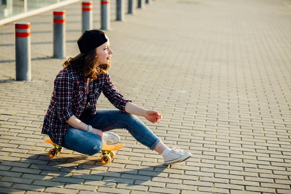 若い魅力的な流行に敏感な女性市にスケート ボードの上に座って格子縞のシャツと白いスニーカーを身に着けています。. — ストック写真