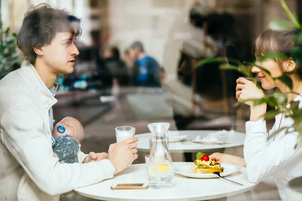 Junge glückliche Familie mit neugeborenem Baby beim Frühstück in der Cafeteria. Papa hält kleinen Sohn, während Mama ihr Frühstück isst. Menschen, gesunder Lebensstil, Familien- und Ernährungskonzept — Stockfoto