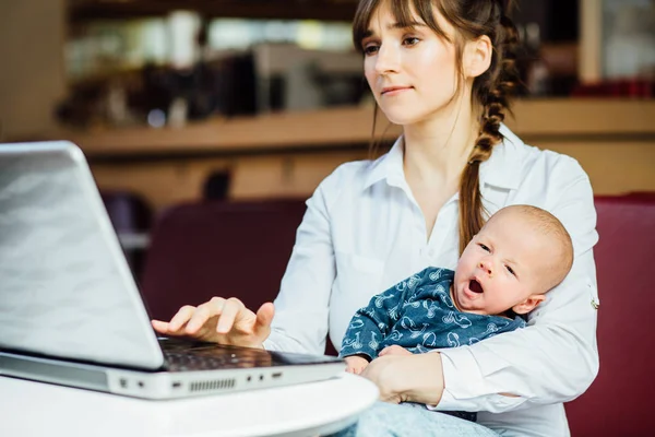 Joven mujer de negocios hermosa y mirando a la computadora portátil mientras está sentada con su bebé en su lugar de trabajo — Foto de Stock