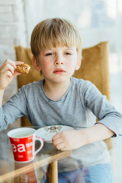 Bonito menino de 5 anos sentado na mesa de vidro e comendo algo com grande gato cinza perto dele . — Fotografia de Stock