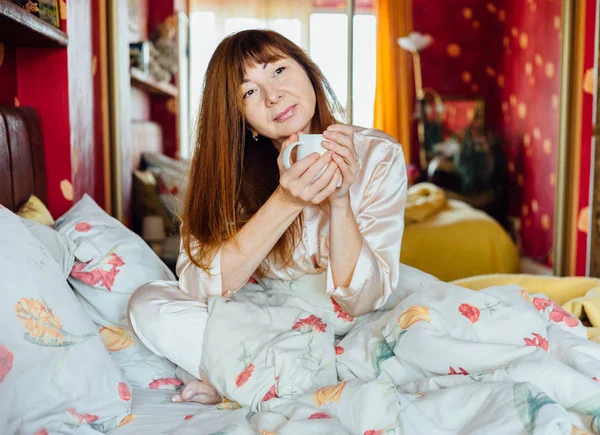 Портрет женщины средних лет с рыжими волосами, сидящей на кровати и держащей белую чашку чая в солнечное утро . — стоковое фото