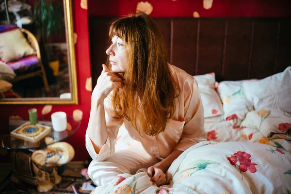 Πορτρέτο της μέσης ηλικίας γυναίκα με τα κόκκινα μαλλιά κοιτάζοντας έξω, ενώ κάθεται στο κρεβάτι το πρωί. — Φωτογραφία Αρχείου
