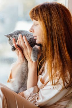 Gri kedi tutarak ve gündoğumu sabah windowshil üzerinde oturan güzel orta yaşlı Kızıl saçlı kadının yakın çekim.