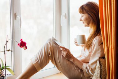 Portrait of positive middle aged woman with red hair sitting on windowsill and holding tablet and white cup of tea in sunny morning. clipart