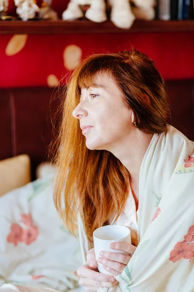 Portrait vertical de charmante femme d'âge moyen aux cheveux roux en vêtements de nuit assis sur le lit avec une tasse de thé le matin ensoleillé . — Photo