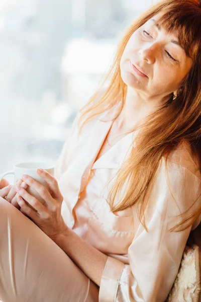 Закрыть портрет женщины средних лет с рыжими волосами, сидящей на подоконнике и держащей белую чашку чая в солнечное утро . — стоковое фото