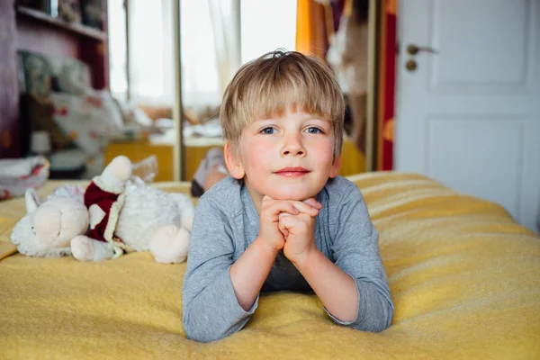 Schattige kleine blond haar jongen op zijn buik op het bed liggen en kijken ernaar uit, grote blauwe ogen jongen af. — Stockfoto