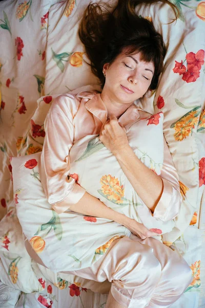 Top view of charming middle aged woman with red brown hair in rose sleepwear higging pillow and slowly sleeping in the bed . — стоковое фото