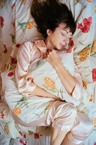 Top view of charming middle aged woman with red brown hair in rose sleepwear higging pillow and slowly sleeping in the bed . — стоковое фото