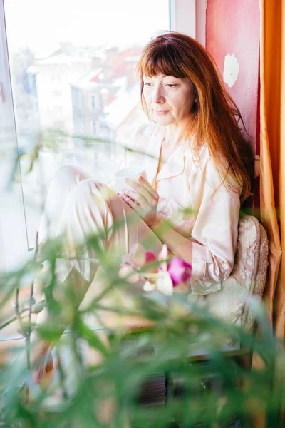 Gülümseyen ve tablet ve beyaz fincan çay güneşli sabah holding pozitif orta yaşlı kadın kahverengi saçlı pencere üzerinde oturan dikey portresi.