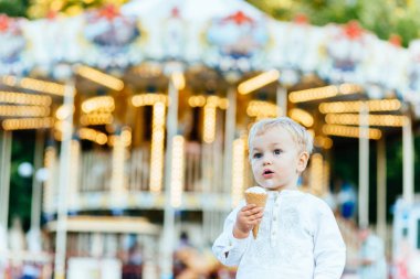 Komik yürümeye başlayan çocuk beyaz gömlek ve Mavi pantolon atlıkarınca önünde bir dondurma yemek