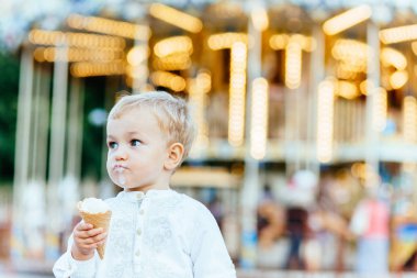 Komik yürümeye başlayan çocuk beyaz gömlek ve Mavi pantolon atlıkarınca önünde bir dondurma yemek