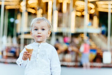 Komik yürümeye başlayan çocuk beyaz gömlek ve Mavi pantolon atlıkarınca önünde bir dondurma yemek
