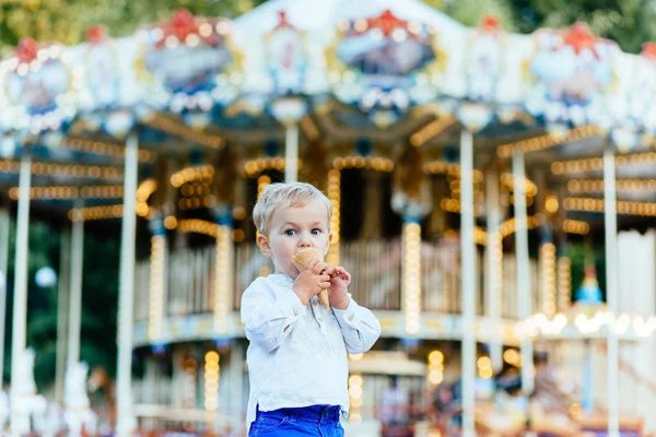 Забавный малыш в белой рубашке и синих штанах ест мороженое перед каруселью — стоковое фото