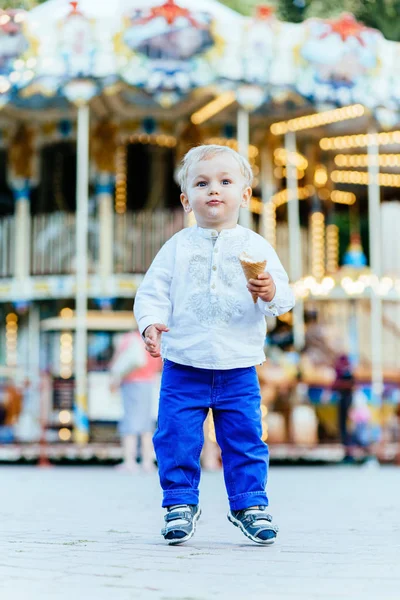 Grappige peuter jongen in wit overhemd en een blauwe broek eten een ijsje voor de carrousel — Stockfoto