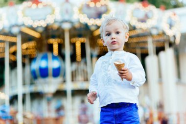 Komik yürümeye başlayan çocuk beyaz gömlek ve Mavi pantolon atlıkarınca önünde bir dondurma yemek