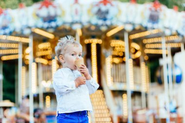 Komik yürümeye başlayan çocuk beyaz gömlek ve Mavi pantolon atlıkarınca önünde bir dondurma yemek