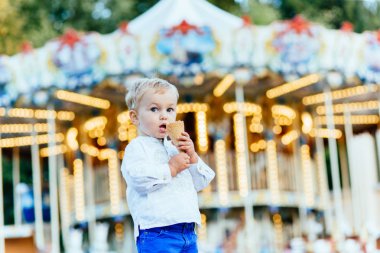 Komik yürümeye başlayan çocuk beyaz gömlek ve Mavi pantolon atlıkarınca önünde bir dondurma yemek