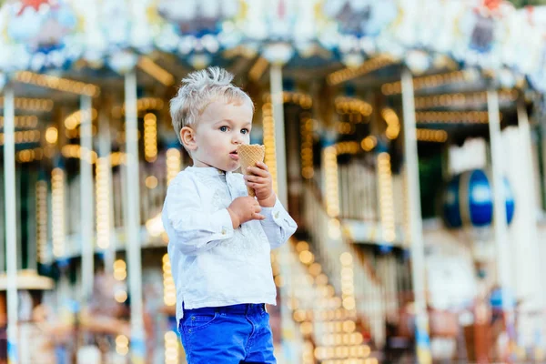 Grappige peuter jongen in wit overhemd en een blauwe broek eten een ijsje voor de carrousel — Stockfoto