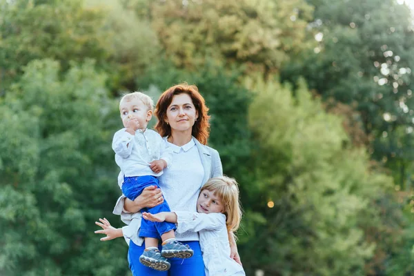 O mamă zâmbitoare fericită, cu doi copii care se distrează împreună pe fundalul copacului verde. Vacanțe de vară și concept de familie . — Fotografie, imagine de stoc