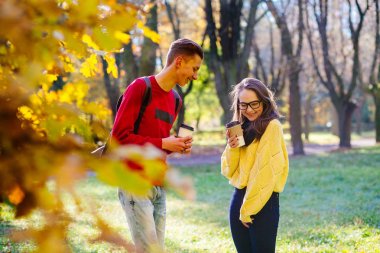 İki üniversite öğrencileri tek kullanımlık Kupası ve gülüyor ve sonbahar bir parkta konuşurken kahve içme. Karışık şüpheli görünümlü erkek ve dişi gençler Güz açık.