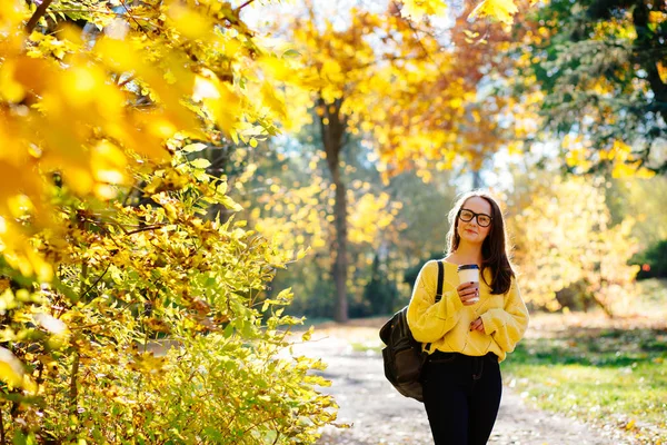 美丽的大学生女孩喝咖啡从一次性杯子在秋季公园 成年女生享受教育 女性穿着黄色毛衣 围巾和眼镜 回到学校 — 图库照片