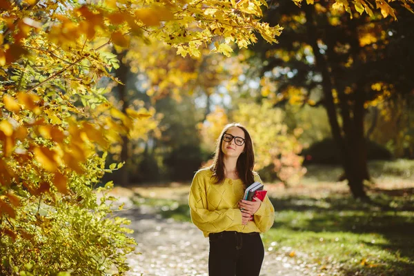 美丽的学生女孩的肖像在秋季公园的书籍 成年女生享受教育 美丽的女性穿着黄色毛衣 围巾和眼镜 回到学校 — 图库照片