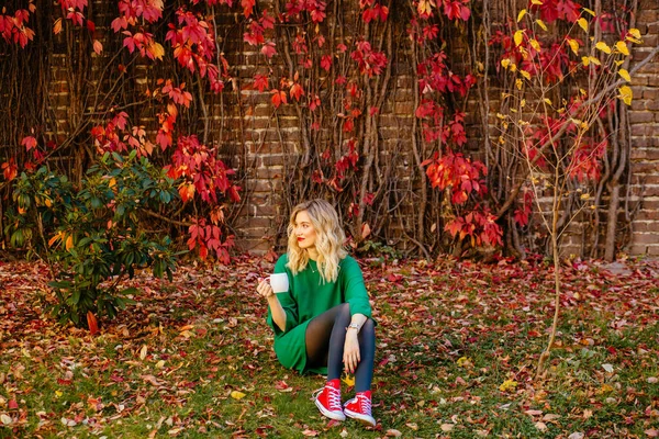 Ρομαντικό όνειρο Ξανθό κορίτσι σε πράσινο πλεκτό πουλόβερ με κούπες στα χέρια στο κόκκινο τοίχο τούβλο με αναρρίχηση σταφύλια στο παρασκήνιο. Φθινοπωρινή Θερμαινόμενη Έννοια. — Φωτογραφία Αρχείου