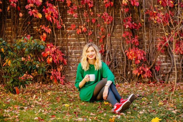 浪漫的梦中的金发姑娘，身穿绿色针织毛衣，手拿着杯子，背景是爬上葡萄的红砖墙。 「秋心热身」概念. — 图库照片