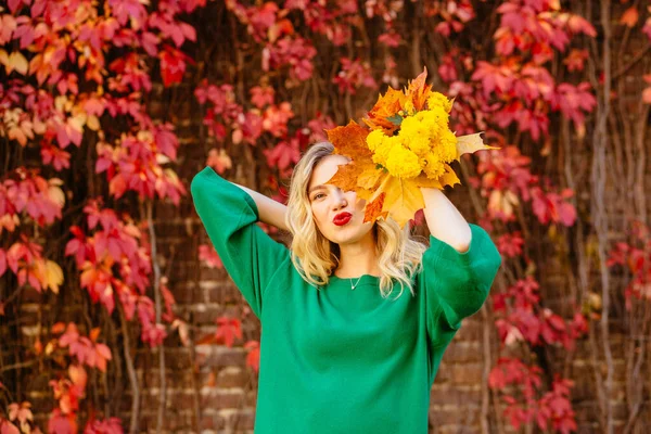 Romantisches verträumtes blondes Mädchen in grünem Strickpullover mit Becher in der Hand, sitzend auf Gras mit bunten Blättern an roter Ziegelwand mit Klettertrauben im Hintergrund. Herbststimmung wärmt Konzept auf. — Stockfoto
