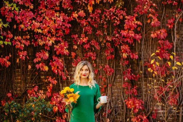Blondynka romantyczna młoda kobieta trzyma kubek z gorącym napojem i żółty bukiet jesienne kwiaty — Zdjęcie stockowe