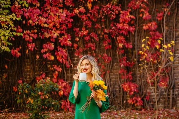 Блондинка романтична молода жінка в зеленому светрі тримає кухоль з гарячим напоєм і жовтими букетиками осінніх квітів на червоній стіні з скелелазінням винограду на фоні. Щасливий спосіб життя, концепція падіння — стокове фото