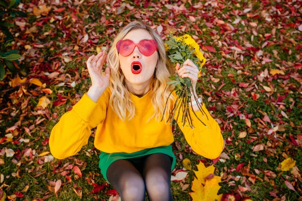 Stylische blonde Hipsterfrau mit gelbem Strickpullover, herzförmiger Sonnenbrille auf Gras sitzend auf herbstlich buntem, hellem Hintergrund, lebendiger Lebensstil der Menschen, entspanntes Urlaubskonzept — Stockfoto