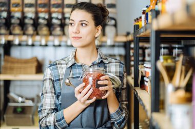 Sıfır atık dükkanı kavramı. Plastiksiz marketin arka planında büyük cam bir kavanoz taşıyan bir kadın. Minimalist vejetaryen kız plastik ambalajsız yiyecekler alıyor..