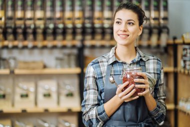 Sıfır atık dükkanı kavramı. Plastiksiz marketin arka planında büyük cam bir kavanoz taşıyan bir kadın. Minimalist vejetaryen kız plastik ambalajsız yiyecekler alıyor..