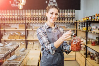 Sıfır Atık Dükkanında Genç Karışık Yarış Kadını Süper Gıda Satıyor. Markette ayakta duran cam şişelerde bir sürü sağlıklı yiyecek var. Plastik Bilinçli Minimalizm Vegan Yaşam Tarzı Konsepti yok. Boşluğu kopyala