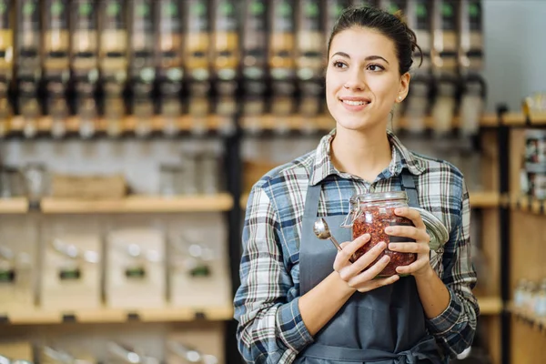 Concept of zero waste shop. Woman holding large glass jar with groceries on interior background of plastic free grocery store. Minimalist vegan style girl buying foods without plastic packaging.