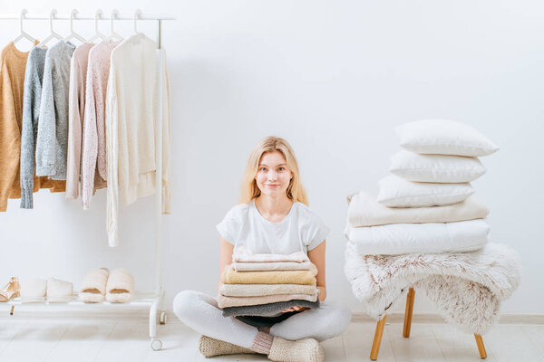 Young woman housewife sitting on the floor holding a stack of pillows, cover plaid on white wall stack tower design in scandinavian style, clothes rack in modern light empty wardrobe room.