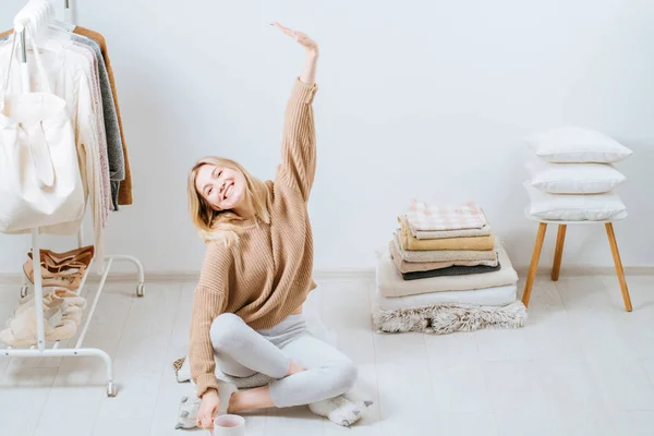 Jonge vrouw zit op de vloer in de buurt van stapel handdoeken kussens, bedek de ruit op witte muur stapel toren naast haar op de stoel. Huis in Scandinavische stijl, kledingrek in moderne lege garderobe. — Stockfoto